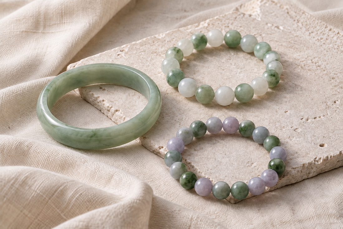Pale green bangle with green, white, and lavender stone bead bracelets arranged on ivory linen and pale stone.