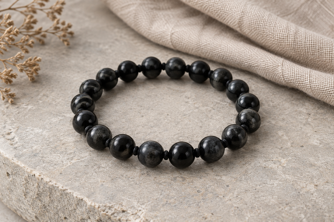 Dark rounded bead bracelet on pale stone beside oatmeal linen and dried stems.